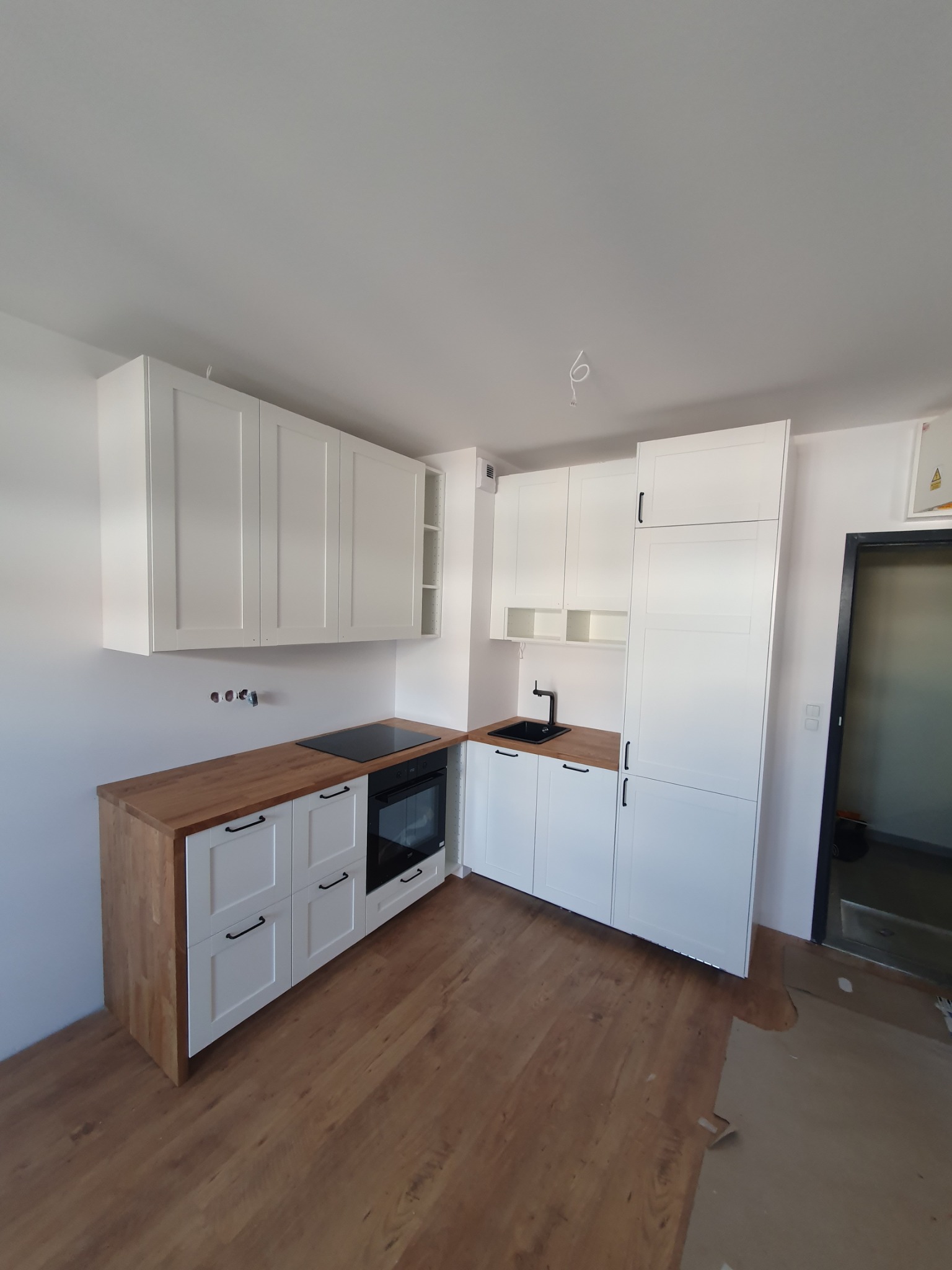 Nowoczesna, jasna kuchnia z drewnianymi blatami i czarnymi uchwytami, jeszcze w trakcie wykańczania, z widocznymi przewodami wystającymi ze ściany i zabezpieczeniami podłogi.