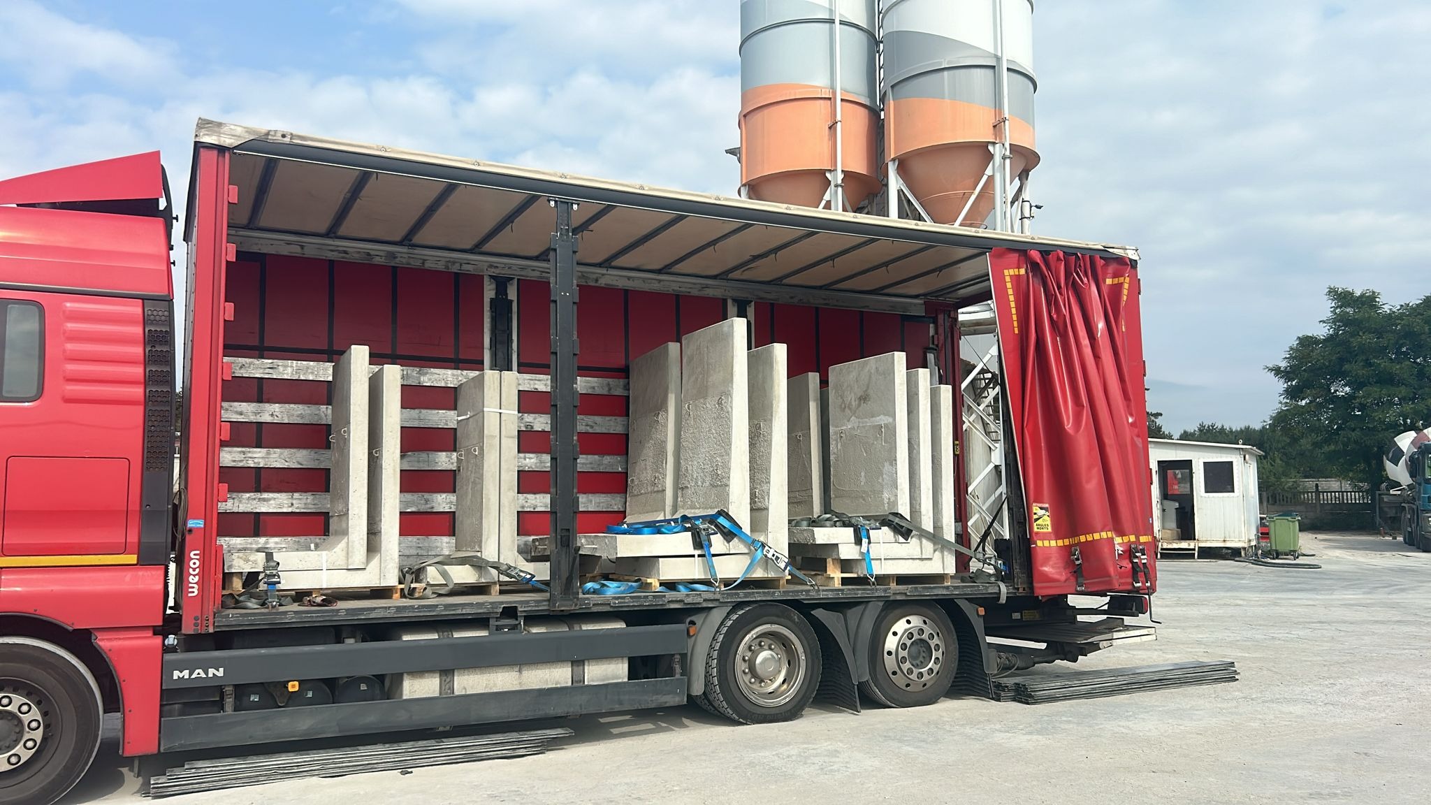 Czerwona ciężarówka z otwartą naczepą załadowana prefabrykatami betonowymi zabezpieczonymi niebieskimi pasami transportowymi, na tle silosy do betonu.