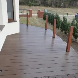 Nowoczesny taras z desek kompozytowych w kolorze ciemnego brązu, z drewnianymi słupkami balustrady, widok na ogród z tujami i samochód w tle.