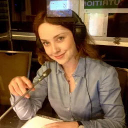 Kobieta z mikrofonem i słuchawkami w studiu nagraniowym, z dokumentem przed sobą i zegarkiem na ręce, uśmiechnięta.