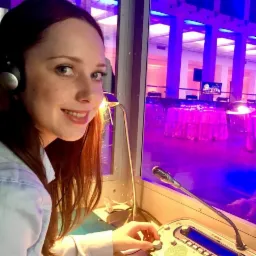 Kobieta w kabinie tłumaczeń symultanicznych z mikrofonem i słuchawkami, uśmiechnięta, z widokiem na salę konferencyjną w tle oświetloną na fioletowo.