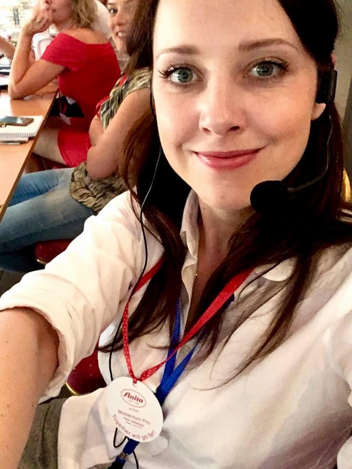 Kobieta z headsetem i identyfikatorem na szyi, uśmiechnięta, w tle inne osoby przy stole.