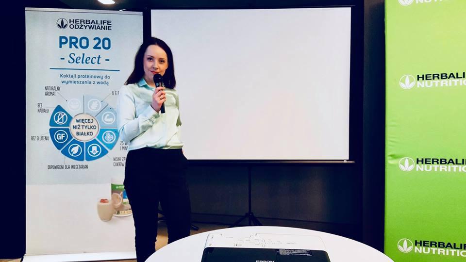 Kobieta z mikrofonem prezentuje produkt Herbalife na tle ekranu projekcyjnego i banerów reklamowych.