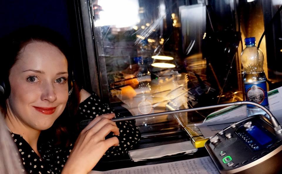 Kobieta w słuchawkach i bluzce w kropki, siedząca w kabinie tłumaczeniowej z mikrofonem i dokumentami, z widocznym odbiciem świateł miasta w szybie.