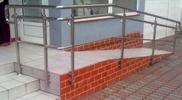 Zewnętrzna rampa dla niepełnosprawnych z metalową balustradą, wykończona czerwoną cegłą, prowadząca do budynku z szarą elewacją.
