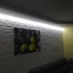 Fragment salonu z dekoracyjną ścianą z białej cegły, podświetloną listwą LED podsufitową i obrazem z motywem kwiatowym.