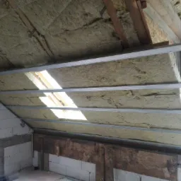 Skośny strych w trakcie ocieplania wełną mineralną, widoczne profile metalowe i fragment okna dachowego wpadającego światła.