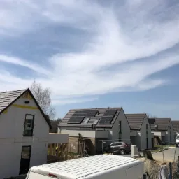 Szereg nowych domów jednorodzinnych w trakcie budowy lub wykończenia, z widocznymi panelami słonecznymi na niektórych dachach i elewacją w stanie surowym na pierwszym planie, pod błękitnym niebem...