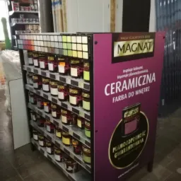 Ekspozytor z farbami Magnat Ceramiczna w sklepie, widoczne puszki z farbami na półkach oraz paleta kolorów na górze, obok drzwi w surowym stanie.