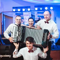 Czteroosobowa grupa muzyczna z akordeonami, w białych koszulach i z muszkami, uśmiechnięta, na scenie z oświetleniem w kolorze niebieskim.