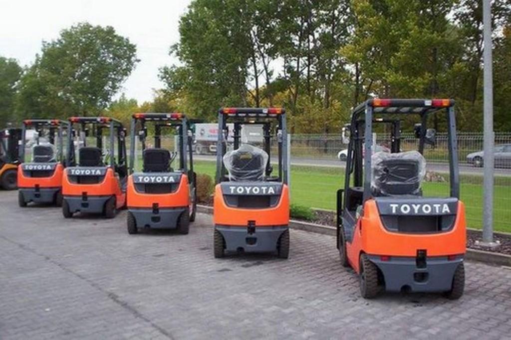 Rząd nowych, pomarańczowo-szarych wózków widłowych marki Toyota, ustawionych na brukowanym placu, z widoczną folią ochronną na siedzeniach, na tle zieleni i ulicy.
