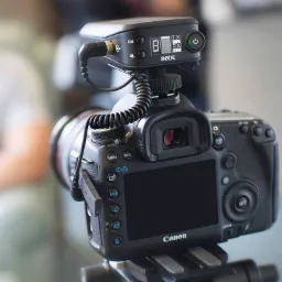 Aparat Canon z podłączonym mikrofonem Rode, widoczny ekran LCD, przyciski funkcyjne i logo Canon, ustawiony na statywie.