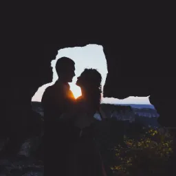 Sylwetki pary młodej w jaskini, z zachodzącym słońcem tworzącym aureolę wokół ich głów i malowniczym krajobrazem w tle.