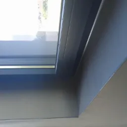 Gładka, szara powierzchnia ściany po nałożeniu tynku maszynowego, z widocznym oknem dachowym i precyzyjnym wykończeniem narożnika.