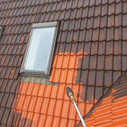 Alpi-GardenMac - Częściowo umyty dach z dachówki ceramicznej, widoczne okno dachowe i lanca myjki ciśnieniowej. Połowa dachu w kolorze ciemnobrązowym, druga w jasnym pomarańczowym.