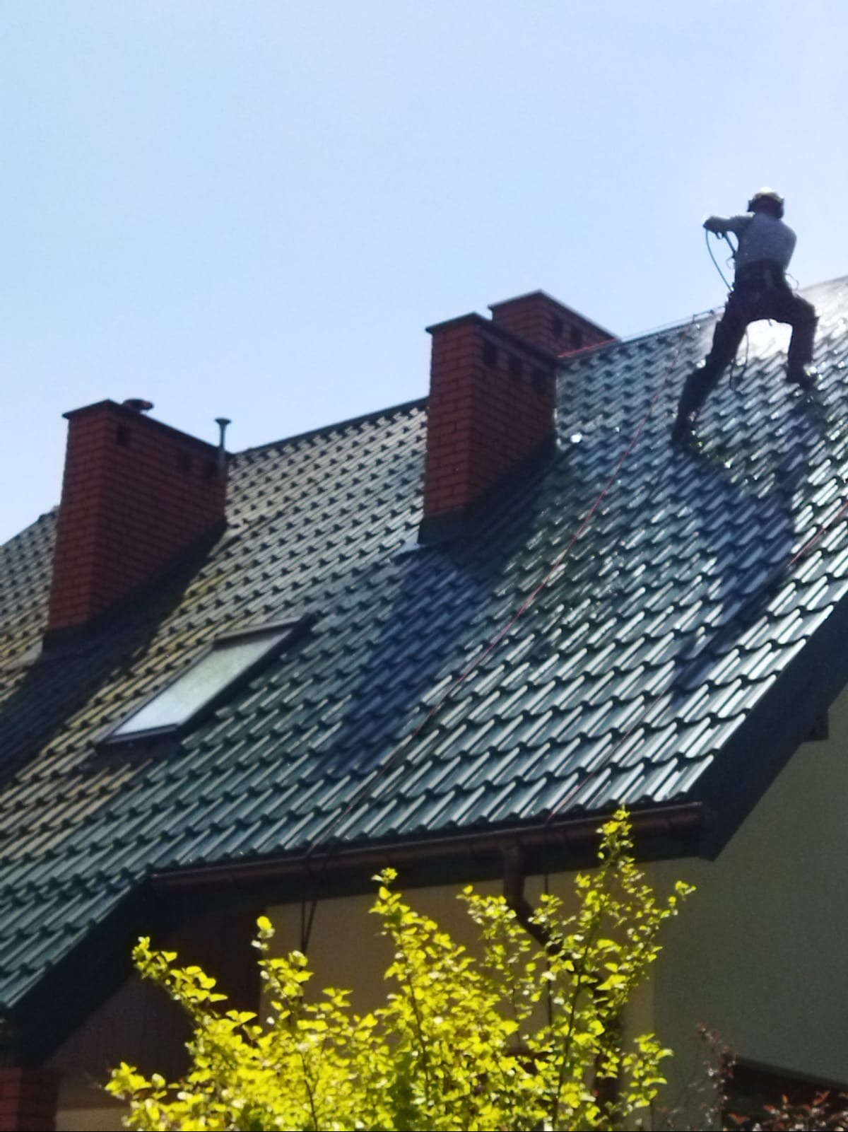 Pracownik w kasku czyści ciemnozielony dach z ceglanymi kominami za pomocą myjki ciśnieniowej. Widok na dom z dołu, z zielonymi krzewami w dole kadru.