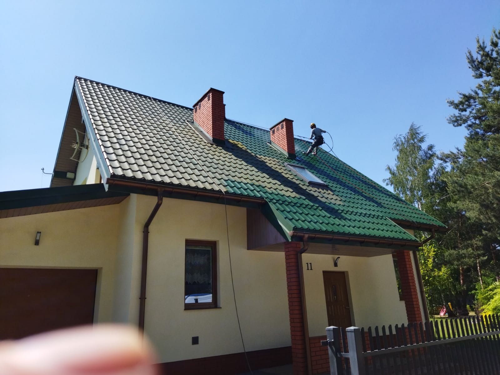 Pracownik w kasku czyści zielony dach domu z ceglanymi kominami za pomocą myjki ciśnieniowej. Widoczne orynnowanie i fragment elewacji budynku.