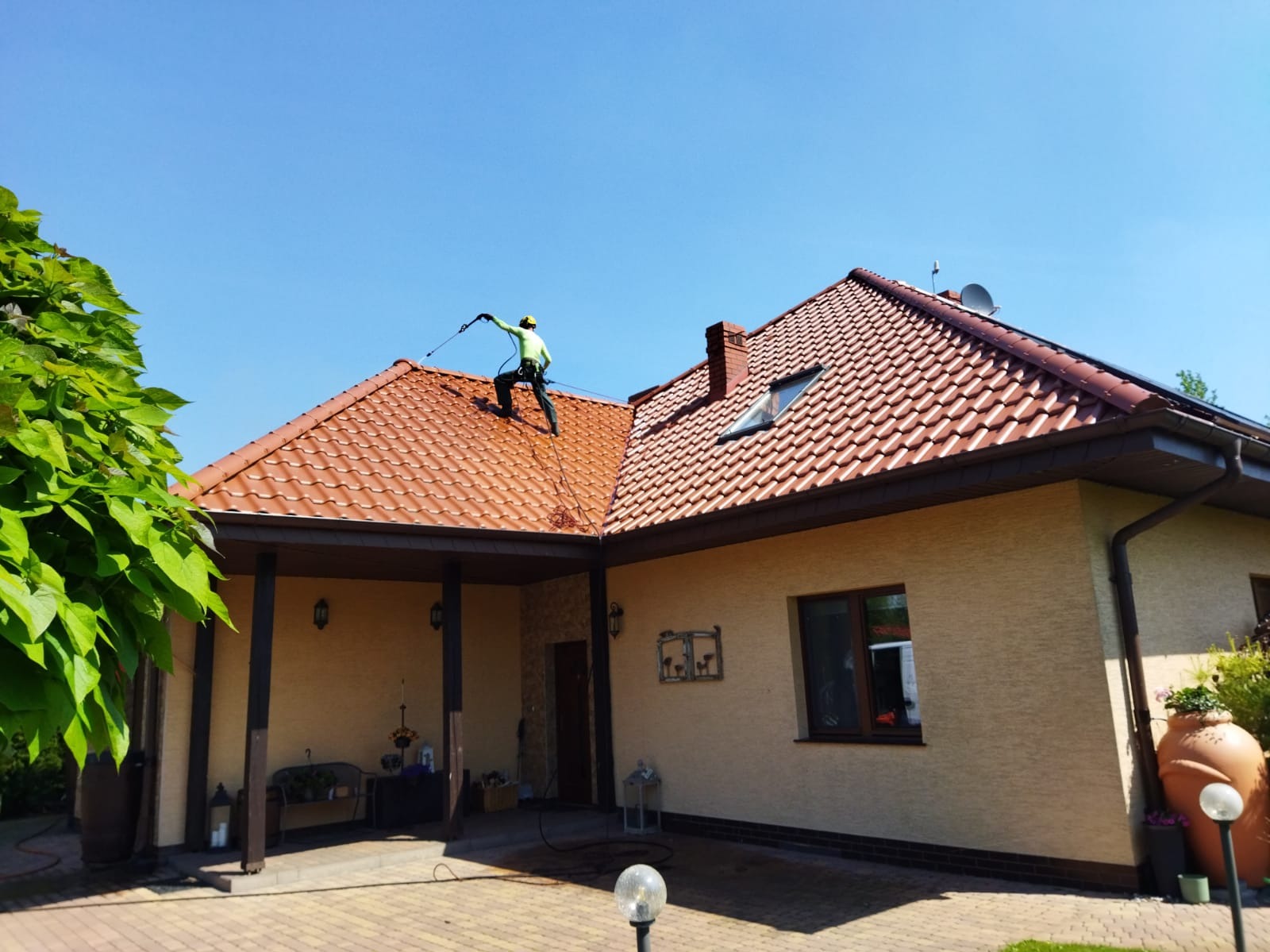 Pracownik w kasku i uprzęży myje czerwoną dachówkę domu jednorodzinnego za pomocą myjki ciśnieniowej. Widoczne zacieki spływającej wody. Błękitne niebo w tle.