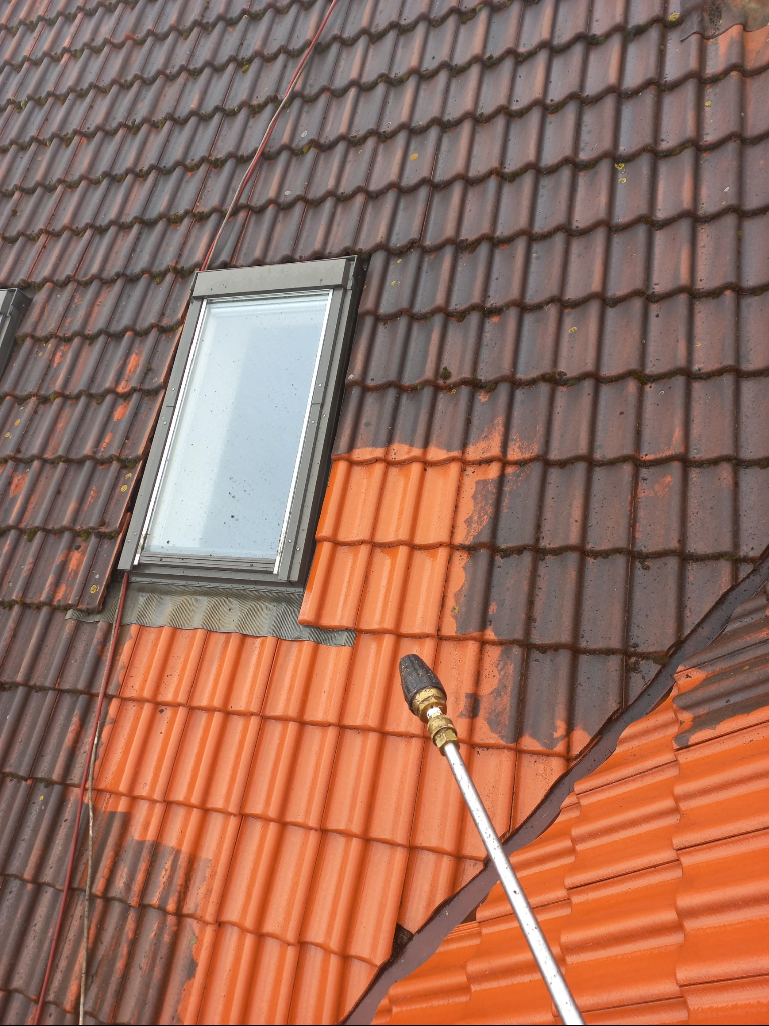 Częściowo umyty dach z dachówki ceramicznej, widoczne okno dachowe i lanca myjki ciśnieniowej. Połowa dachu w kolorze ciemnobrązowym, druga w jasnym pomarańczowym.