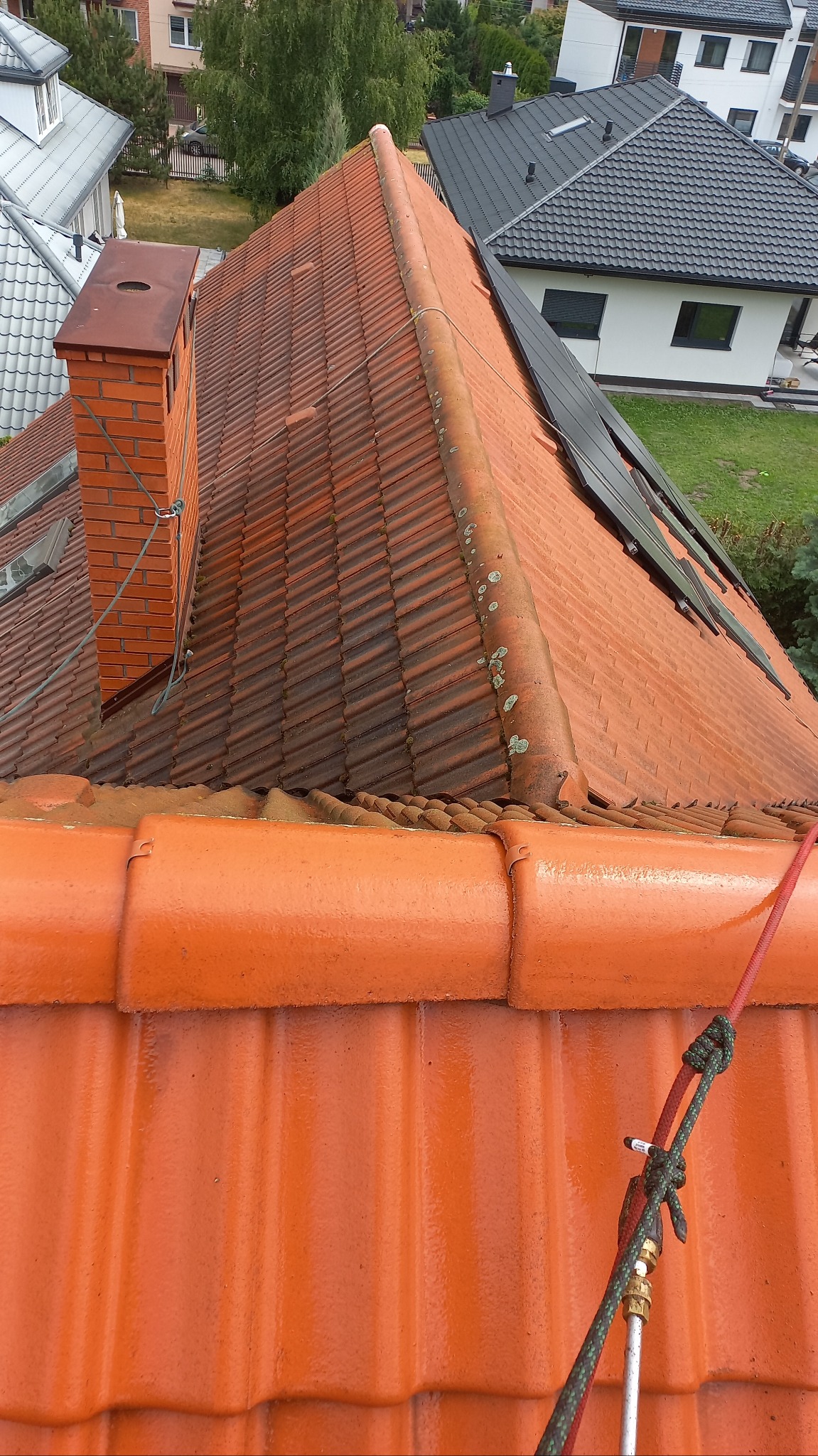 Czerwona dachówka z widocznym zabrudzeniem i mchem, z bliska, z linami zabezpieczającymi, sugerująca prace na wysokości związane z czyszczeniem lub konserwacją dachu.