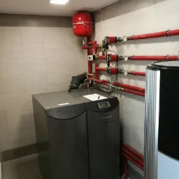 Szary piec grzewczy marki HEIMAR z czerwonym zbiornikiem wyrównawczym i instalacją hydrauliczną z rur w kolorze czerwonym w pomieszczeniu z jasnymi płytkami.