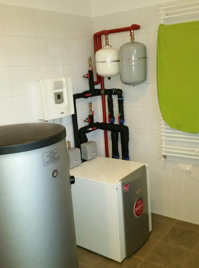 Kompaktowa kotłownia w pomieszczeniu z białymi płytkami, widoczne zbiorniki wyrównawcze, zasobnik ciepłej wody i kocioł grzewczy Wolf BWS-1.