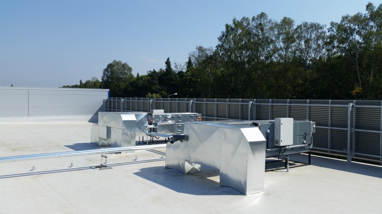 Instalacja wentylacyjna z metalowymi przewodami na dachu budynku przemysłowego, widoczne elementy montażowe i tło z drzewami.