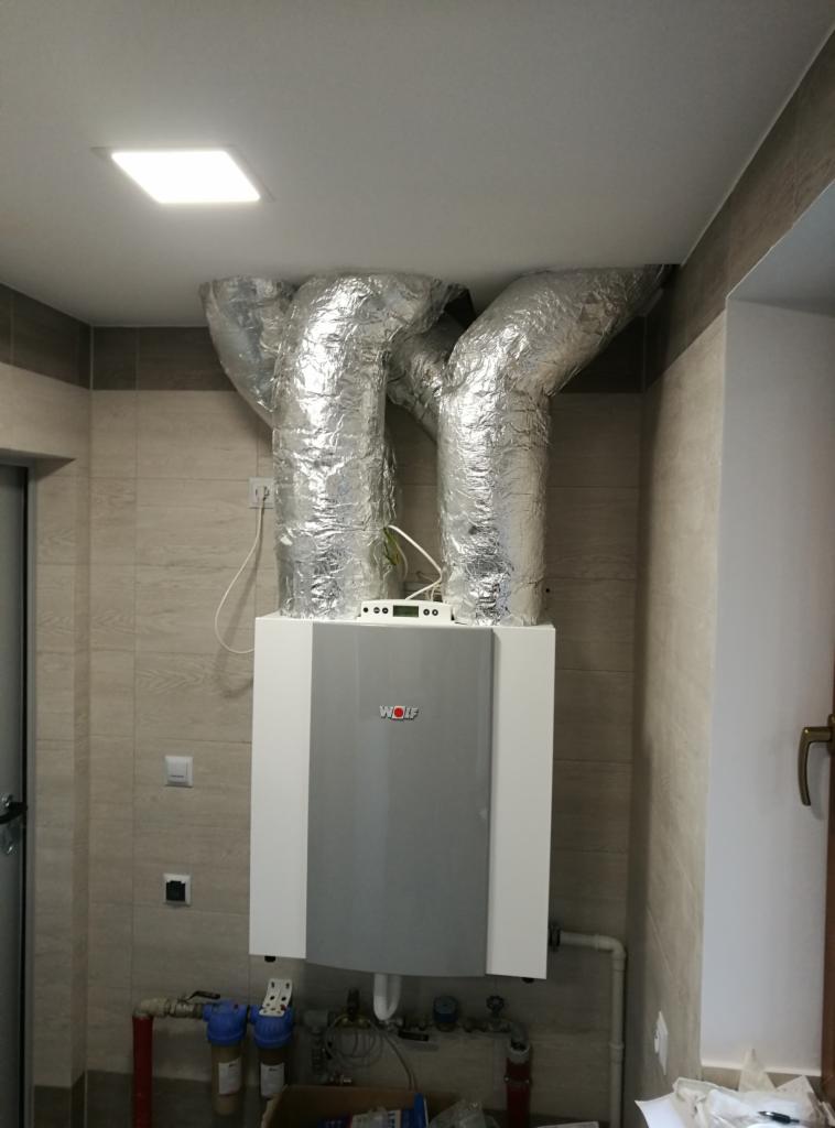 Biało-szary piec gazowy marki Wolf z podłączonymi, izolowanymi przewodami wentylacyjnymi w pomieszczeniu z beżowymi płytkami.