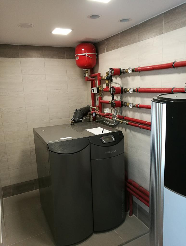 Szary piec grzewczy marki HEIMAR z czerwonym zbiornikiem wyrównawczym i instalacją hydrauliczną z rur w kolorze czerwonym w pomieszczeniu z jasnymi płytkami.