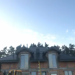 Nowy, grafitowy dach z blachodachówki na budynku w trakcie budowy, widoczne okna dachowe i ceglana elewacja, w tle las iglasty, w kadrze fragment koparki.