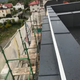 Widok z góry na fragment dachu z blachy na rąbek stojący, z widocznymi śladami deszczu, obok rusztowanie z siatką ochronną i widok na zabudowę jednorodzinną w tle.