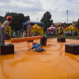 Dzieci w pomarańczowych kaskach bawią się na dużej, pomarańczowej, dmuchanej platformie z czarnymi kostkami i dmuchanym walcem, w tle kolorowe namioty i grupa ludzi.