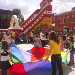 Dzieci bawiące się kolorową chustą animacyjną na tle dmuchanej zjeżdżalni i miejskiej zabudowy.