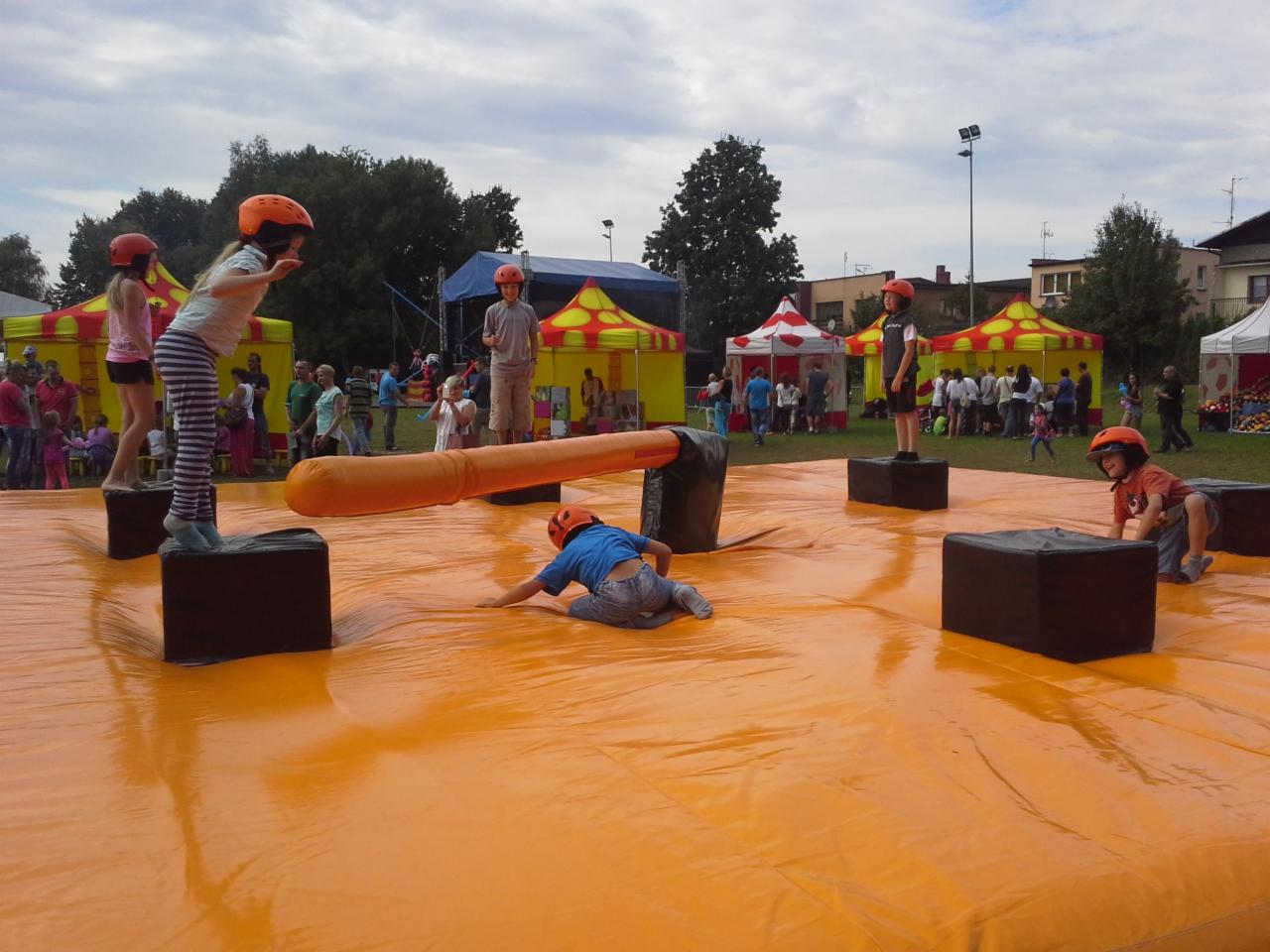 Dzieci w pomarańczowych kaskach bawią się na dużej, pomarańczowej, dmuchanej platformie z czarnymi kostkami i dmuchanym walcem, w tle kolorowe namioty i grupa ludzi.