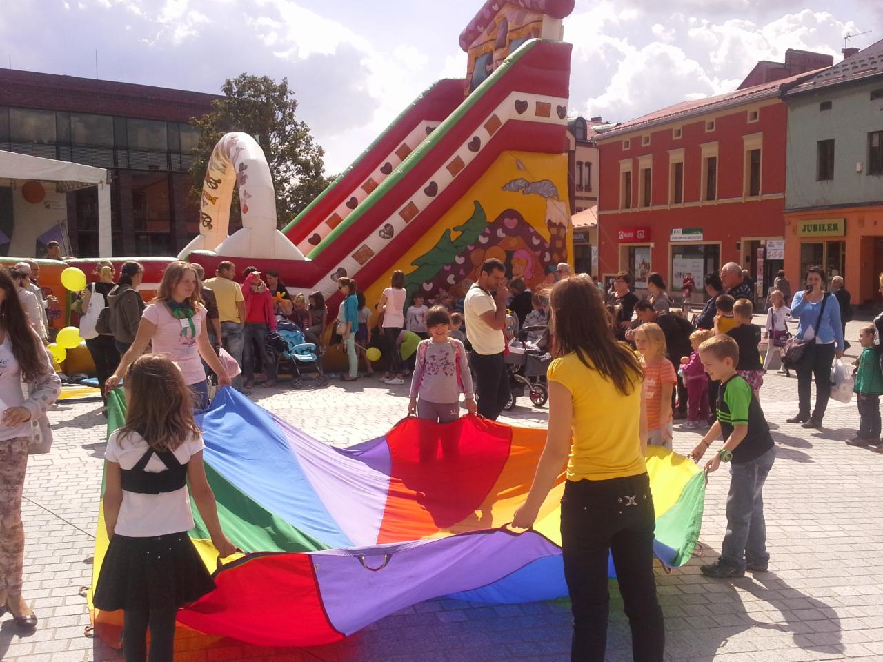 Dzieci bawiące się kolorową chustą animacyjną na tle dmuchanej zjeżdżalni i miejskiej zabudowy.