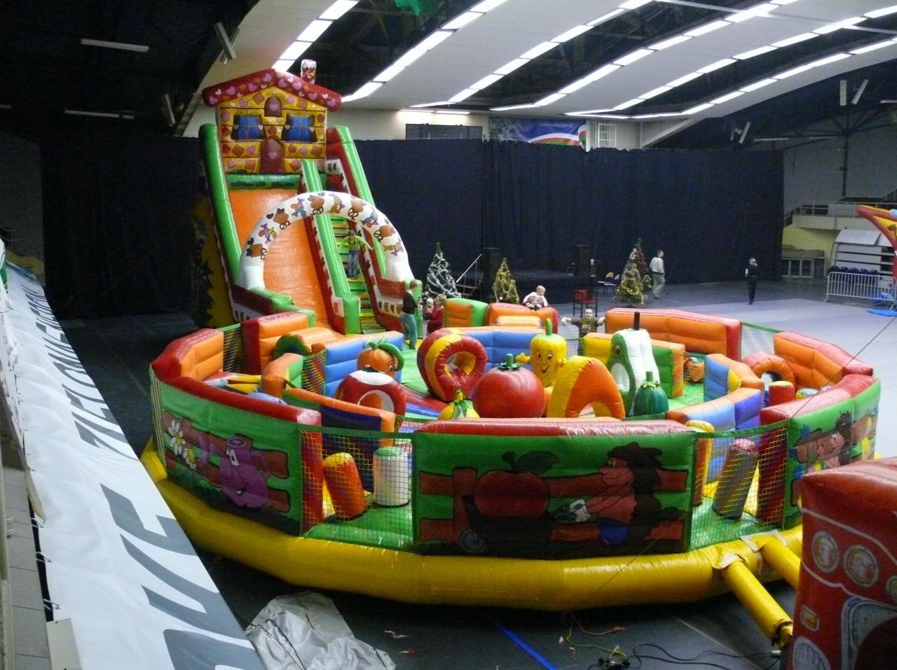 Duży, kolorowy dmuchany plac zabaw z motywem domku z piernika i owoców, ustawiony w hali widowiskowej z czarnym tłem sceny.