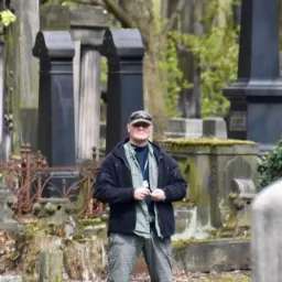 Mężczyzna w ubraniu taktycznym, stojący na cmentarzu, prawdopodobnie detektyw podczas śledztwa.