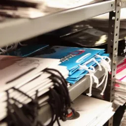 Półki magazynowe z ułożonymi stosami papierowych toreb z uchwytami sznurkowymi w różnych kolorach: białym, czarnym, niebieskim, białym z różowym akcentem, niektóre z nadrukowanym logo.