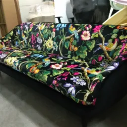 Sofa z czarną tapicerką w motywy kwiatowe i ptasie, widoczna z boku w pomieszczeniu magazynowym.