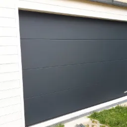 Nowoczesna, segmentowa brama garażowa w kolorze antracytowym z poziomymi przetłoczeniami, zamontowana w białej ramie budynku z drewnianą elewacją. Widoczny fragment trawnika z kratkami trawnikowymi...