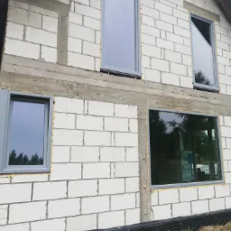 Surowy stan elewacji domu z betonu komórkowego z zamontowanymi szarymi oknami z widoczną pianką montażową wokół ram okiennych.