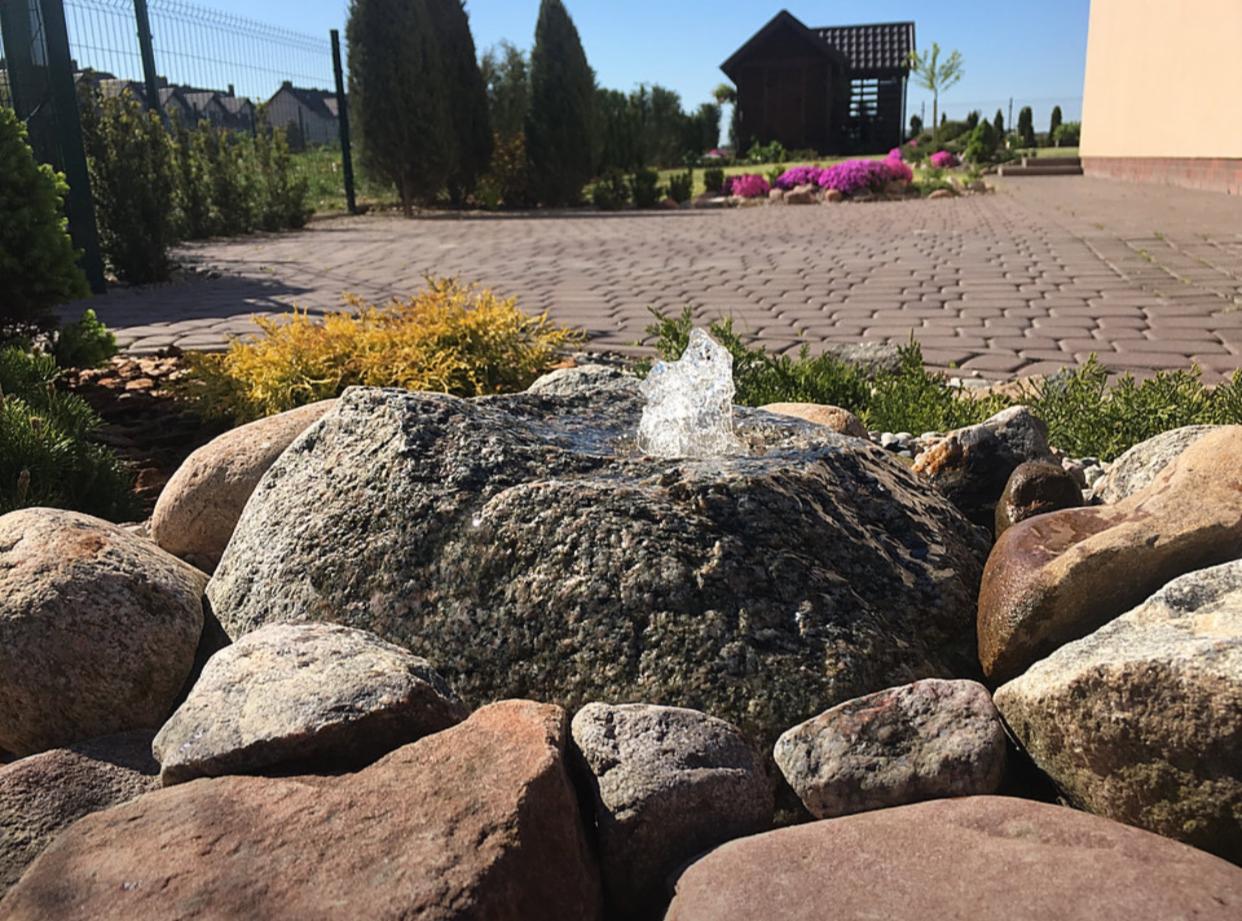 Ogród z fontanną skalną, gdzie woda tryska z dużego, szarego kamienia otoczonego mniejszymi, brązowymi głazami, na tle brukowanej ścieżki i altany.