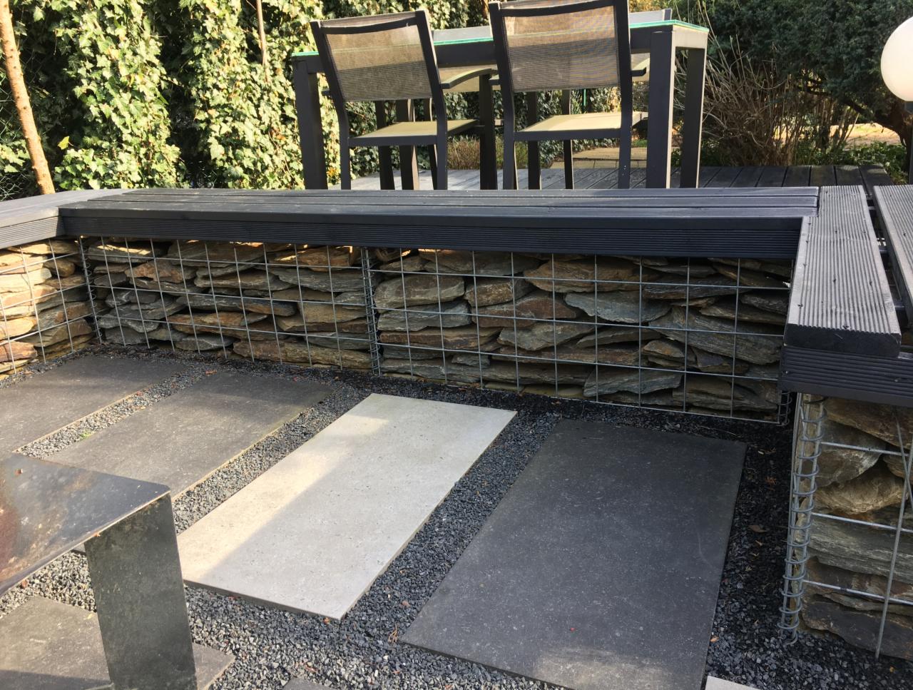 Gabionowa ławka z kamiennym wypełnieniem i drewnianym siedziskiem, otoczona płytami chodnikowymi z drobnym żwirem, w tle stół z krzesłami i zielona ściana roślin.
