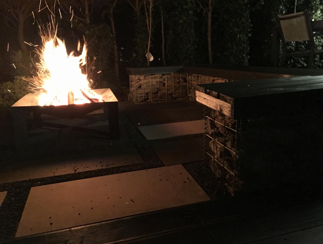 Wieczorny widok na palenisko ogrodowe z płomieniami i iskrami, otoczone siedziskami z gabionów i płytami chodnikowymi.