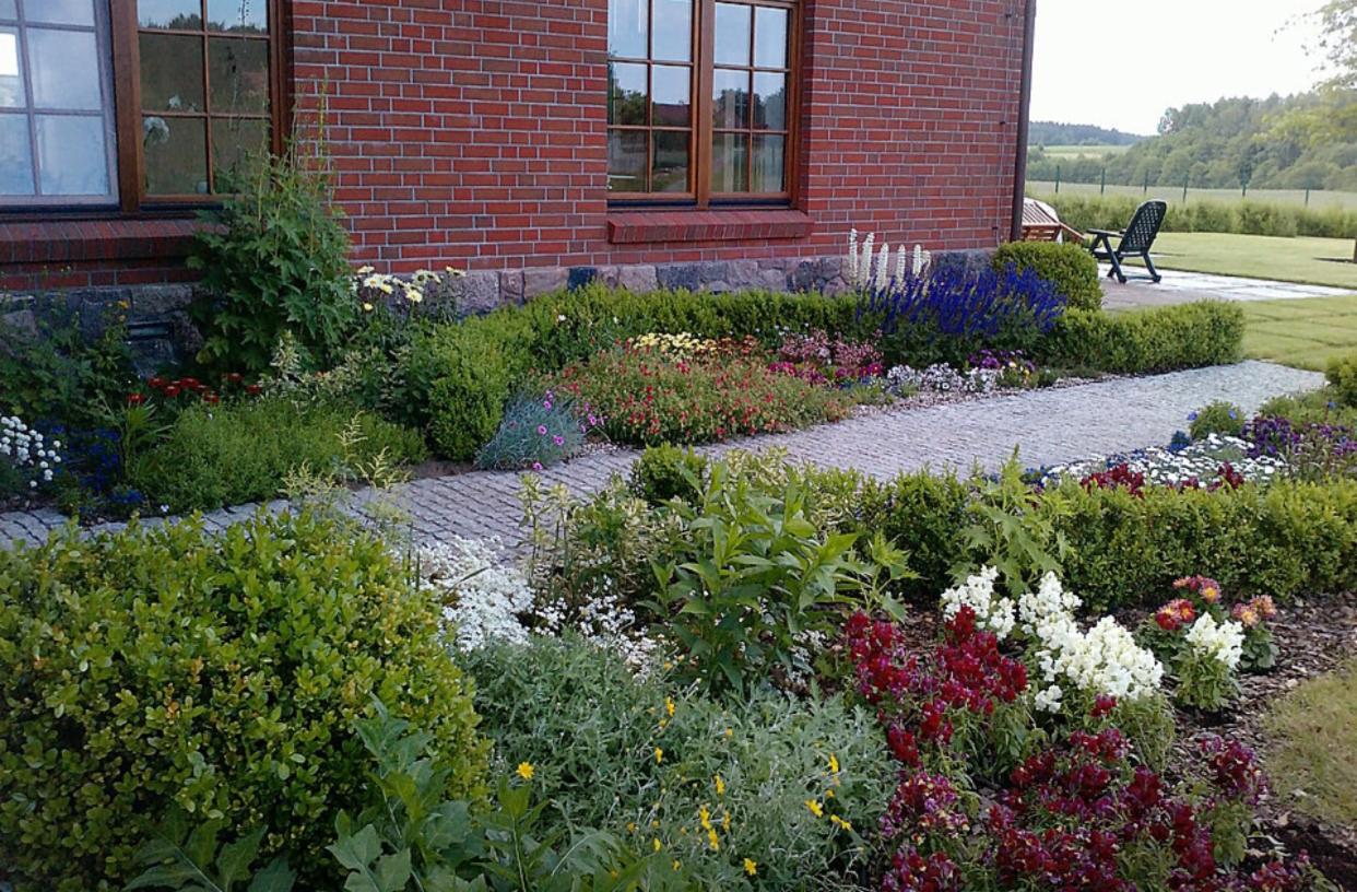 Kaskadowy ogród przydomowy z brukowaną ścieżką, niskimi żywopłotami, kwitnącymi rabatami i ceglanym budynkiem w tle.