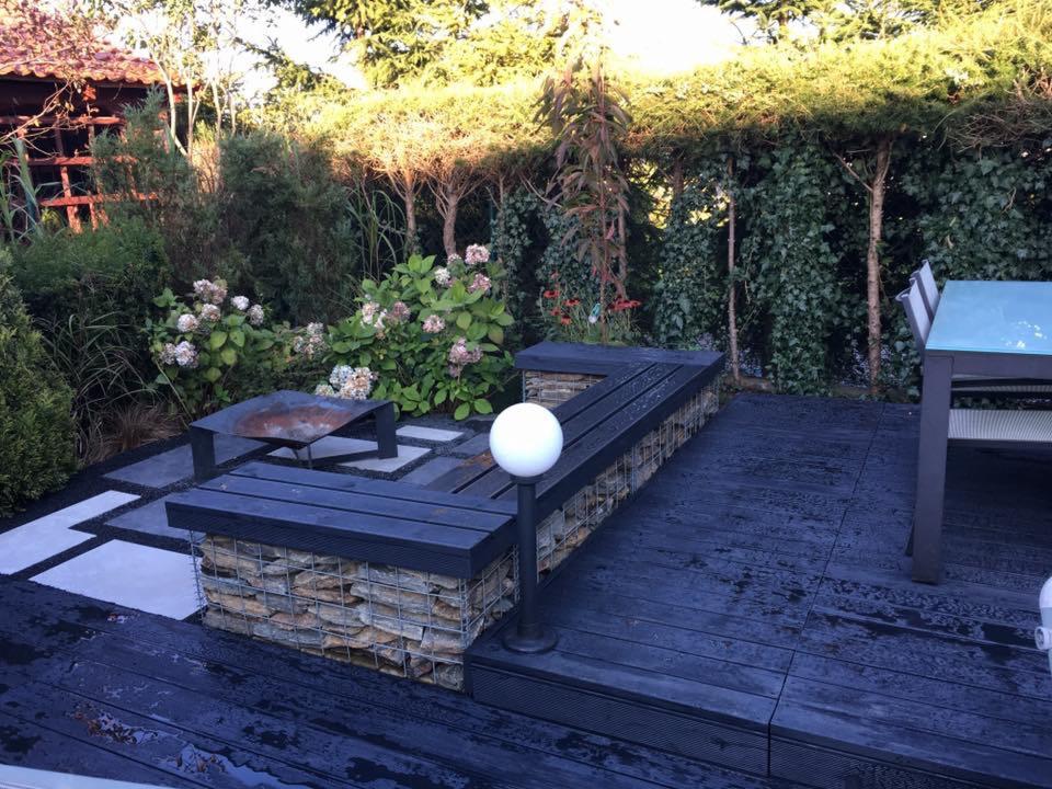 Nowoczesny taras z ciemnego drewna z gabionową ławką, metalowym stolikiem kawowym i kulistą lampą, otoczony zielenią i jasnymi płytami chodnikowymi.