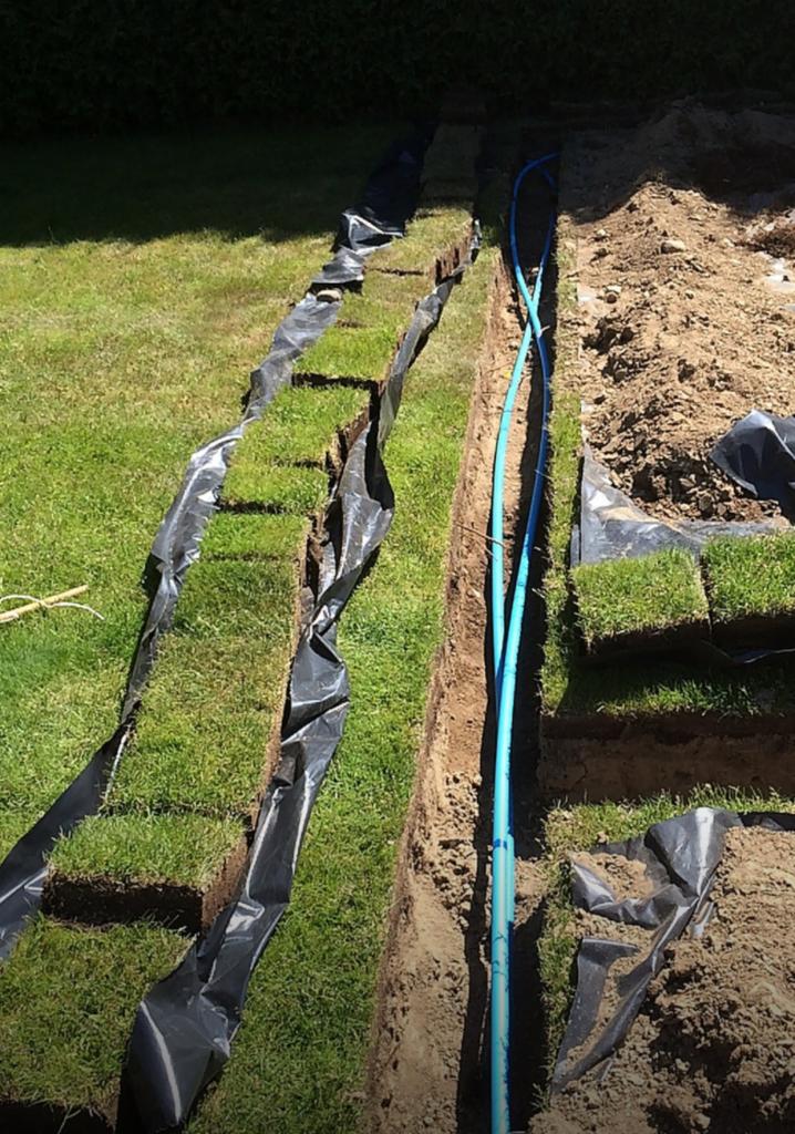 Wykopane rowy w trawniku z ułożonymi kwadratami darni po jednej stronie i niebieskimi rurami w rowie po drugiej stronie, folia ochronna częściowo rozłożona.