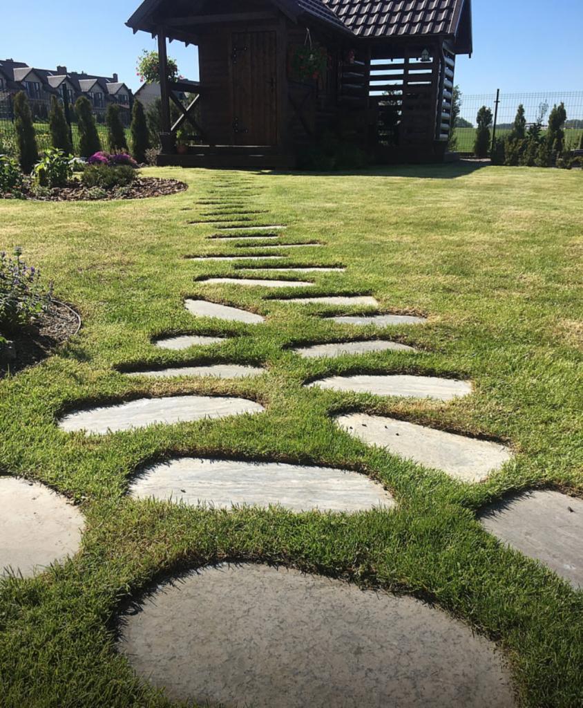 Ścieżka z nieregularnych płyt kamiennych prowadząca do drewnianej altany w zadbanym ogrodzie z tujami.