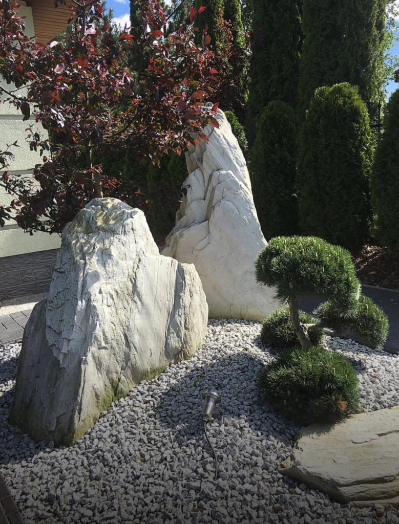 Ogród skalny z dużymi, jasnymi głazami, ozdobną sosną bonsai i podświetleniem punktowym, wysypany drobnym żwirem.