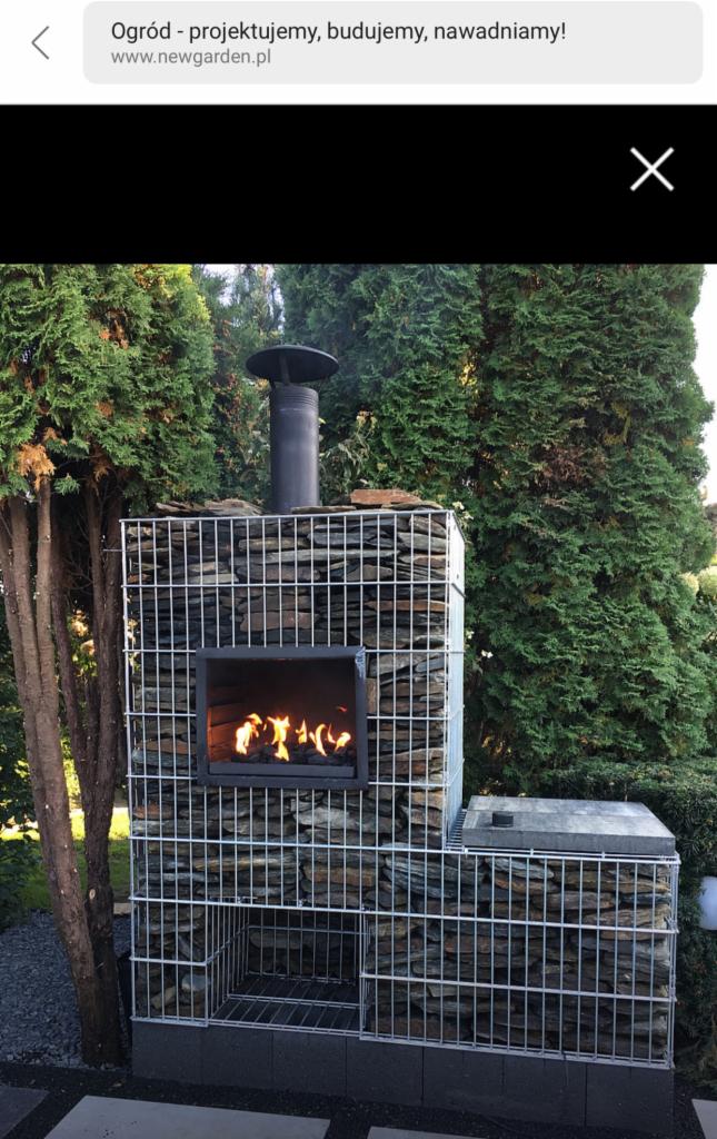 Ogrzewacz ogrodowy z paleniskiem obudowany gabionami wypełnionymi kamieniami, z kominem, otoczony zielonymi drzewami.